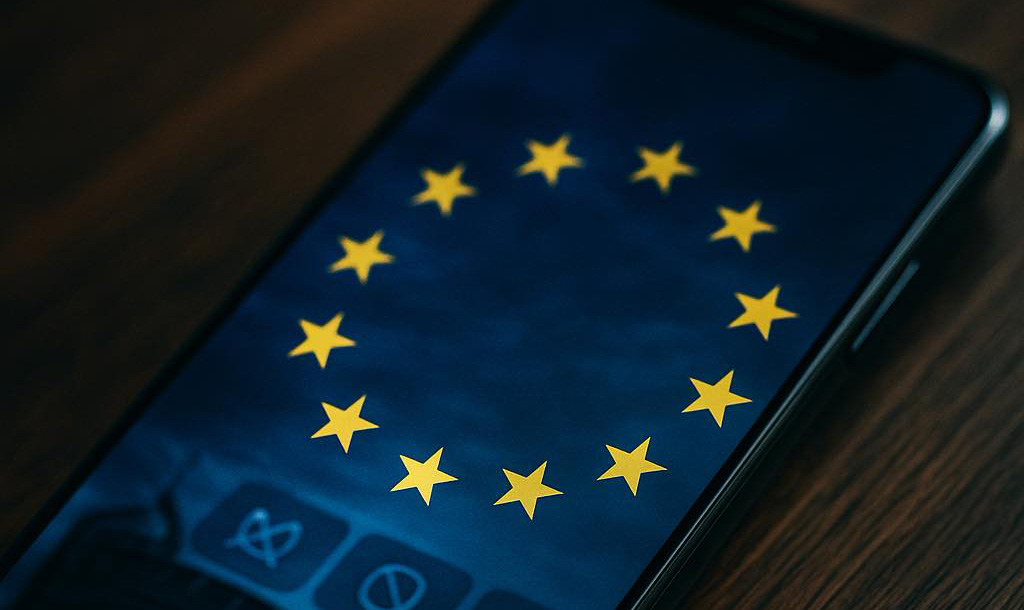 Smartphone op houten tafel met fel verlicht EU-vlag-scherm – visuele hint dat Europese wetgeving invloed heeft op mobiele telefoons.