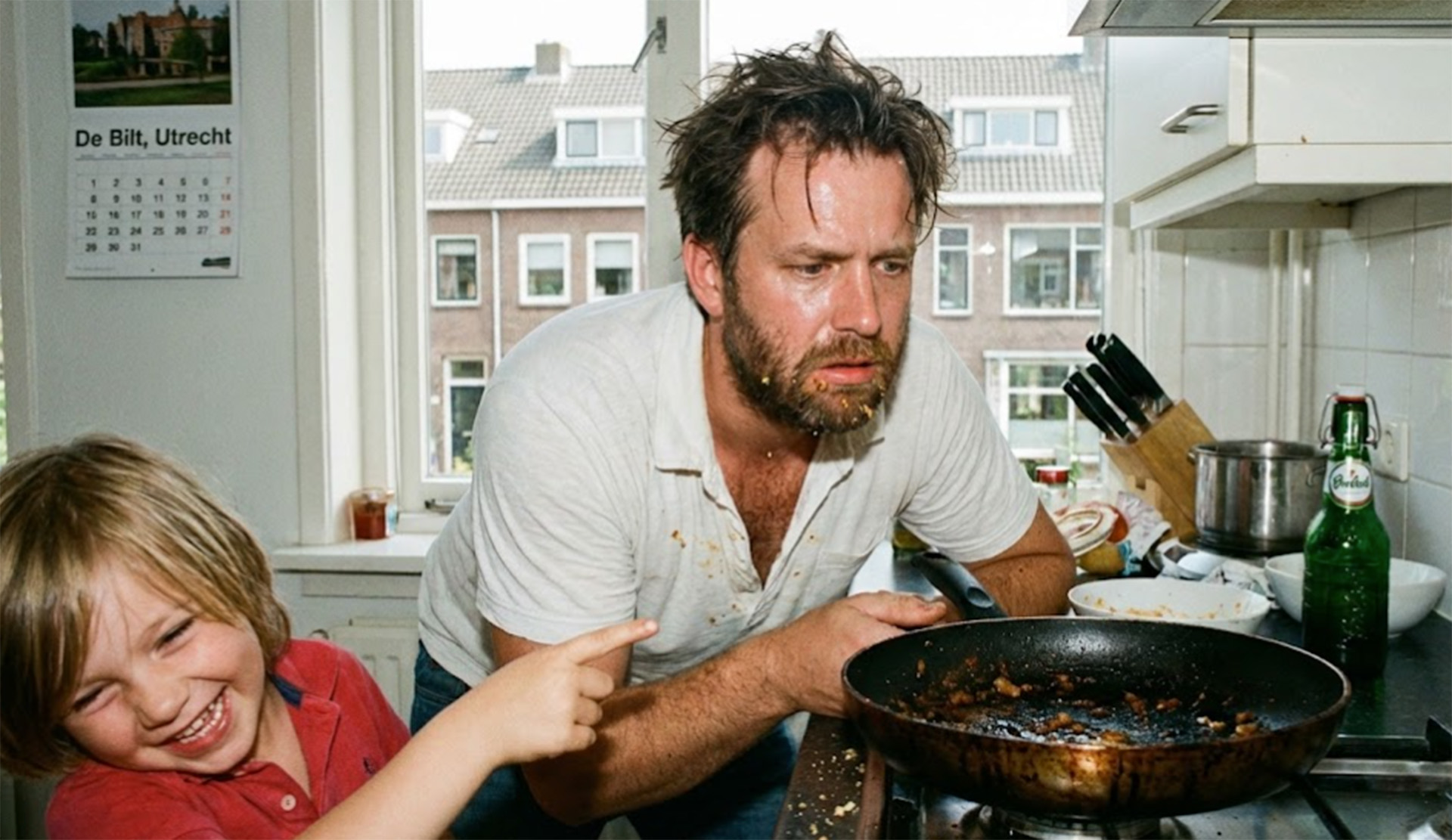 Een gefrustreerde man kijkt naar zijn aangekoekte pan. Naast hem zit een kind te lachen.